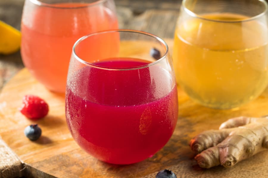 Different colored glasses of kombucha on table with fruit and ginger